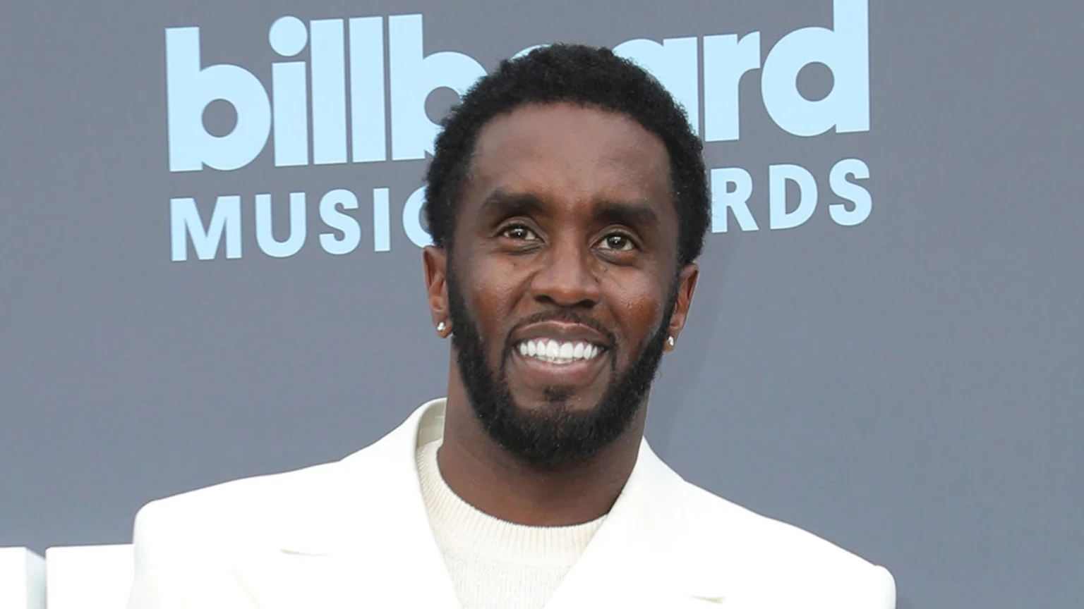 Sean 'Diddy' Combs smiles at the camera, wearing a white suit jacket over a light sweater, with a "Billboard Music Awards" sign in the background.