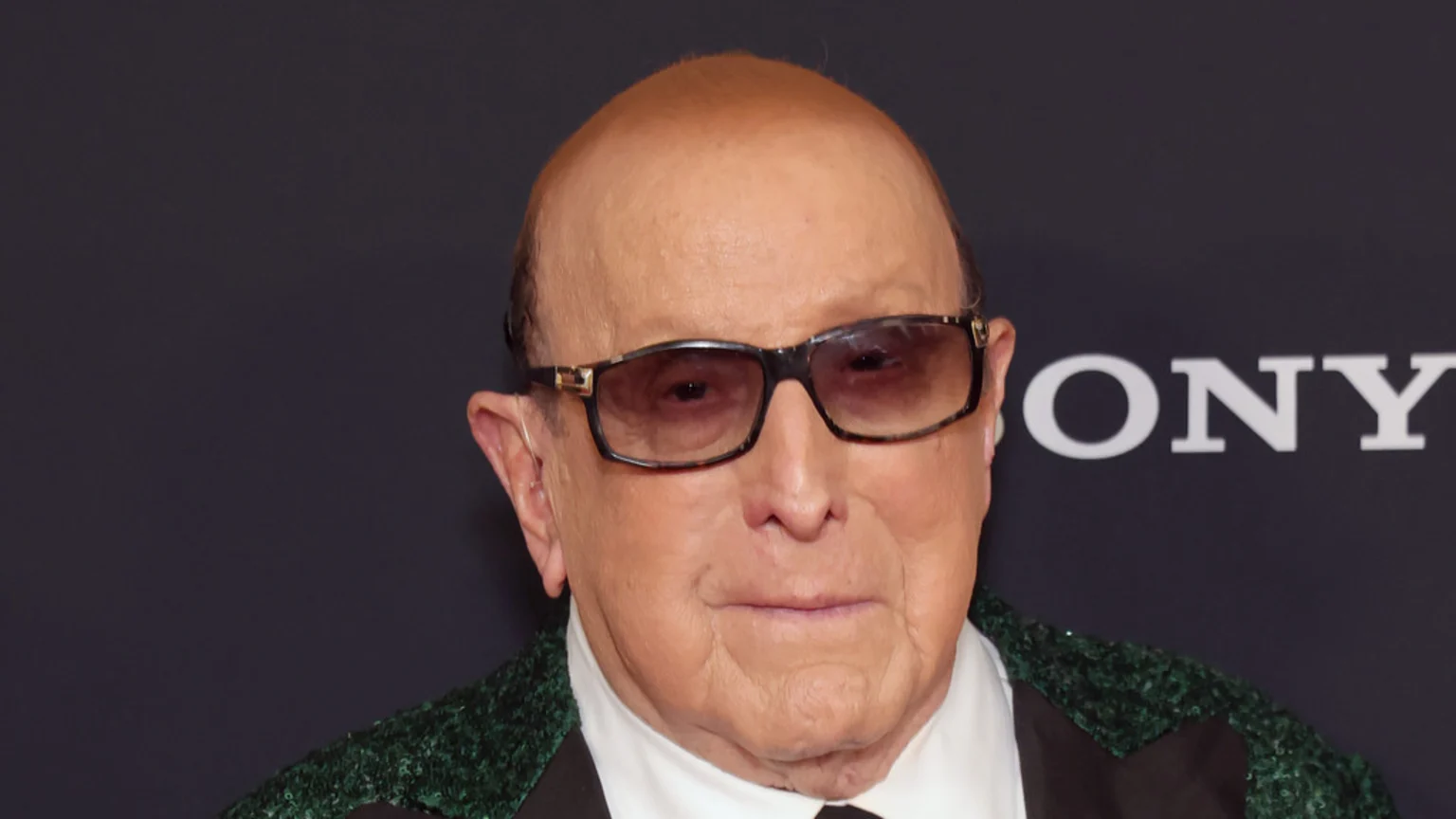A close-up shows an older, bald man with dark sunglasses and a visible hearing aid, wearing a white shirt and a green sequined jacket. The "SONY" logo is in the background.