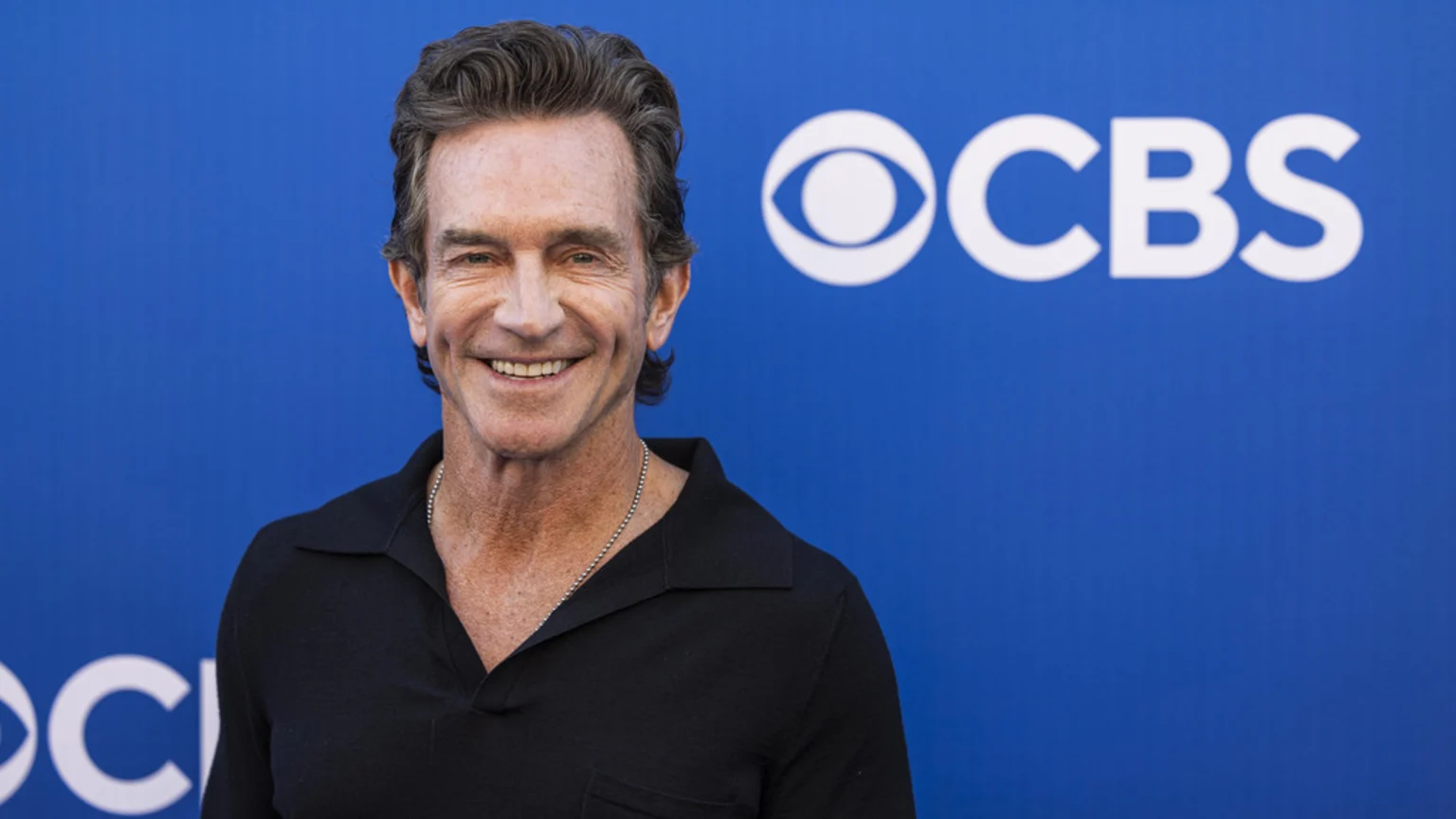 A smiling, fair-skinned man with wavy grey-brown hair and a black polo shirt wears a silver chain necklace in front of a blue background with the white CBS logo.