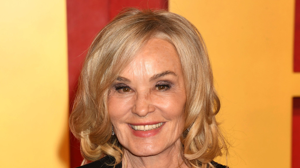 A smiling Jessica Lange, a woman with shoulder-length blonde hair and dark eye makeup, poses for a close-up shot against a blurred yellow and red background.