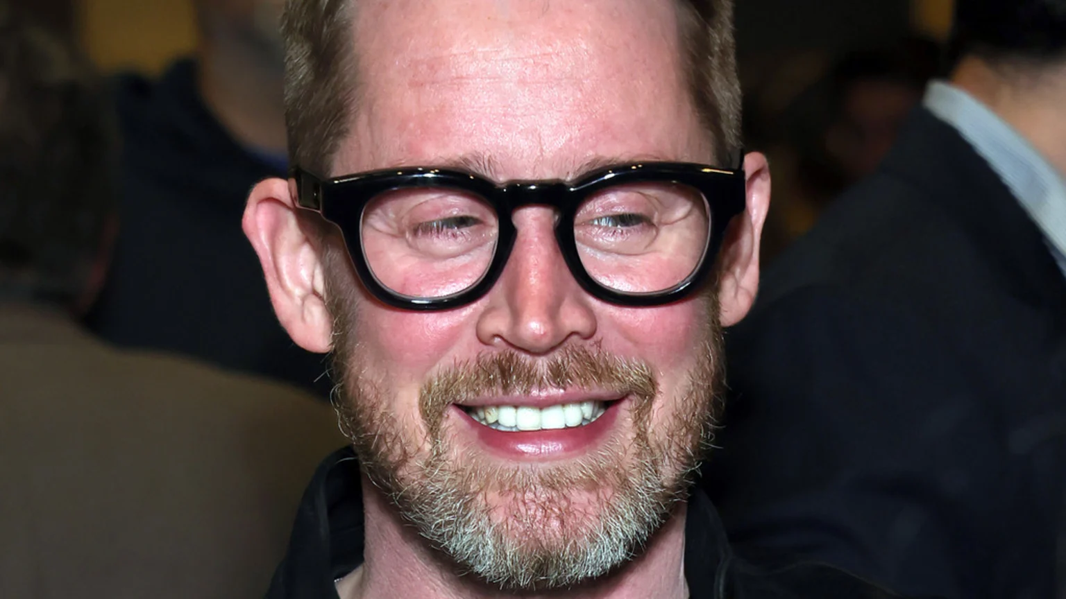 A smiling man with a reddish complexion, a beard, and black glasses looks forward, showing his teeth against a dark, blurry background.