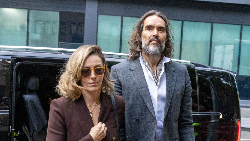Russell Brand, with long hair and a beard, wearing a gray suit, stands next to a woman with blonde hair, sunglasses, and a brown blazer, next to a black vehicle.
