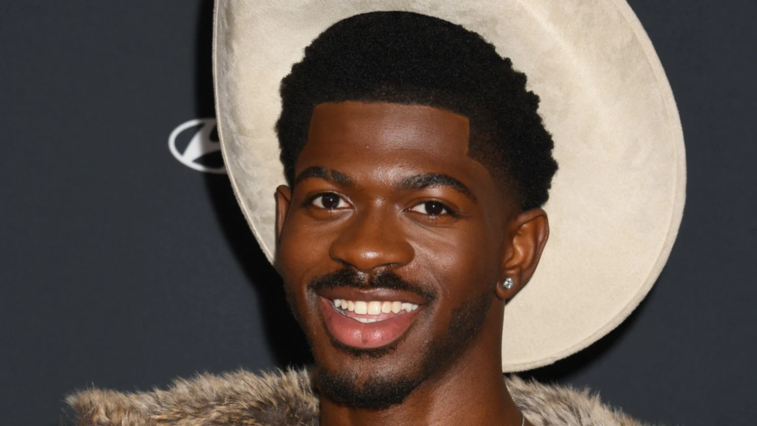 A close-up of Lil Nas X smiling, wearing a light cowboy hat and a fur-trimmed outfit, with a partial white logo visible in the dark background.