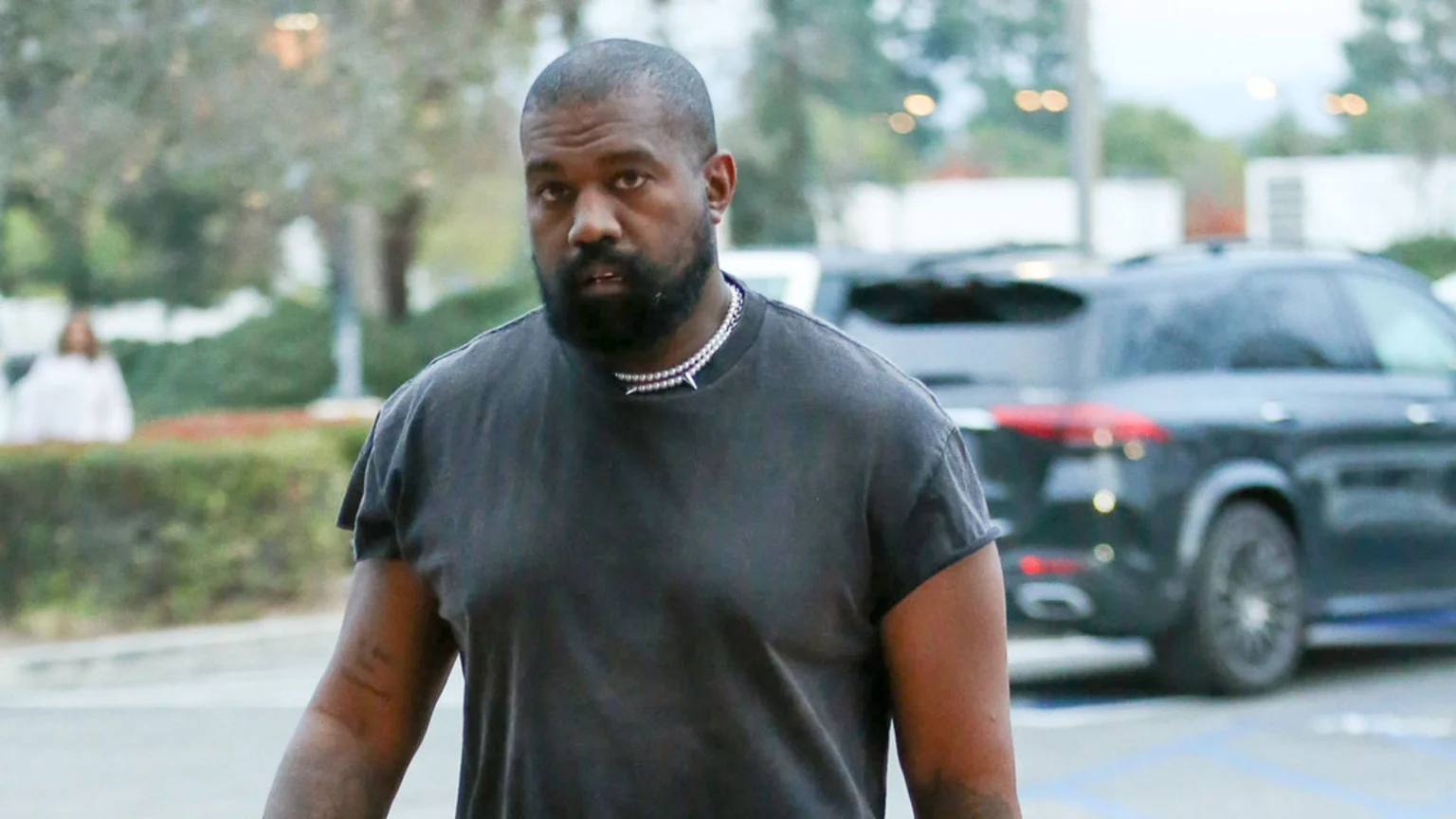 Kanye West, with a beard and short hair, wears a dark grey t-shirt and a silver studded necklace, looking directly forward with a neutral expression.
