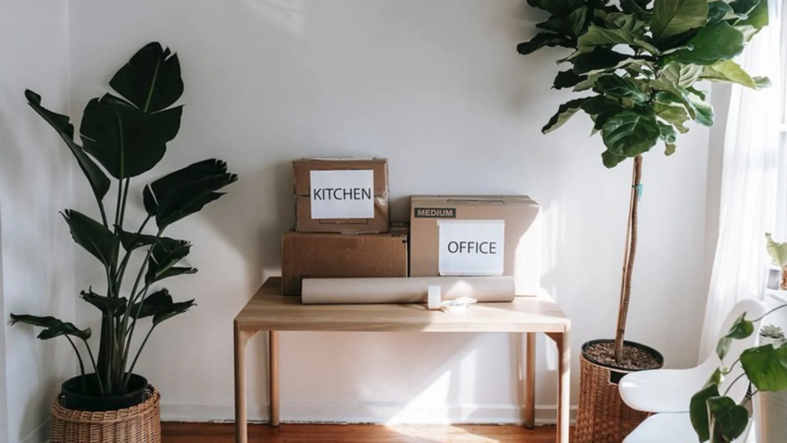 A room with white walls, a wooden table holding two cardboard boxes labeled "KITCHEN" and "OFFICE", packing paper and tape, and two large potted plants in wicker baskets.
