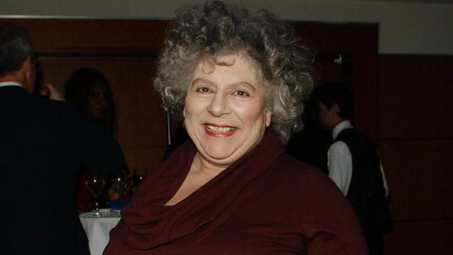 A smiling older woman with curly grey hair and red lipstick wears a dark red top, looking at the camera, with blurry figures and wine glasses visible in the dark background.