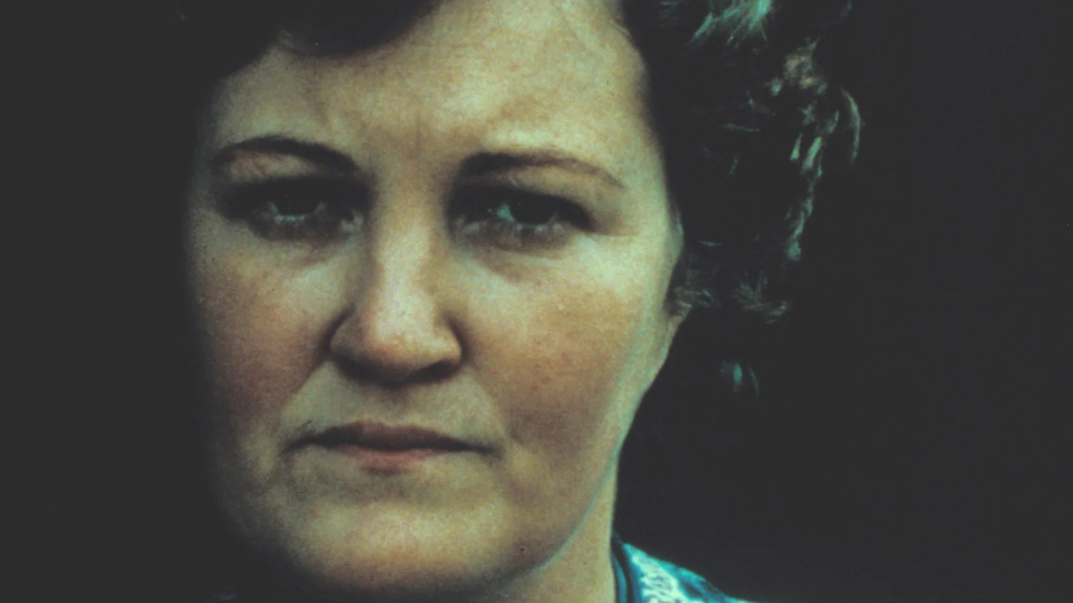 A close-up portrait shows a light-skinned woman with dark, curly hair looking directly at the viewer with a neutral expression, against a dark background, wearing a patterned blue and white top.