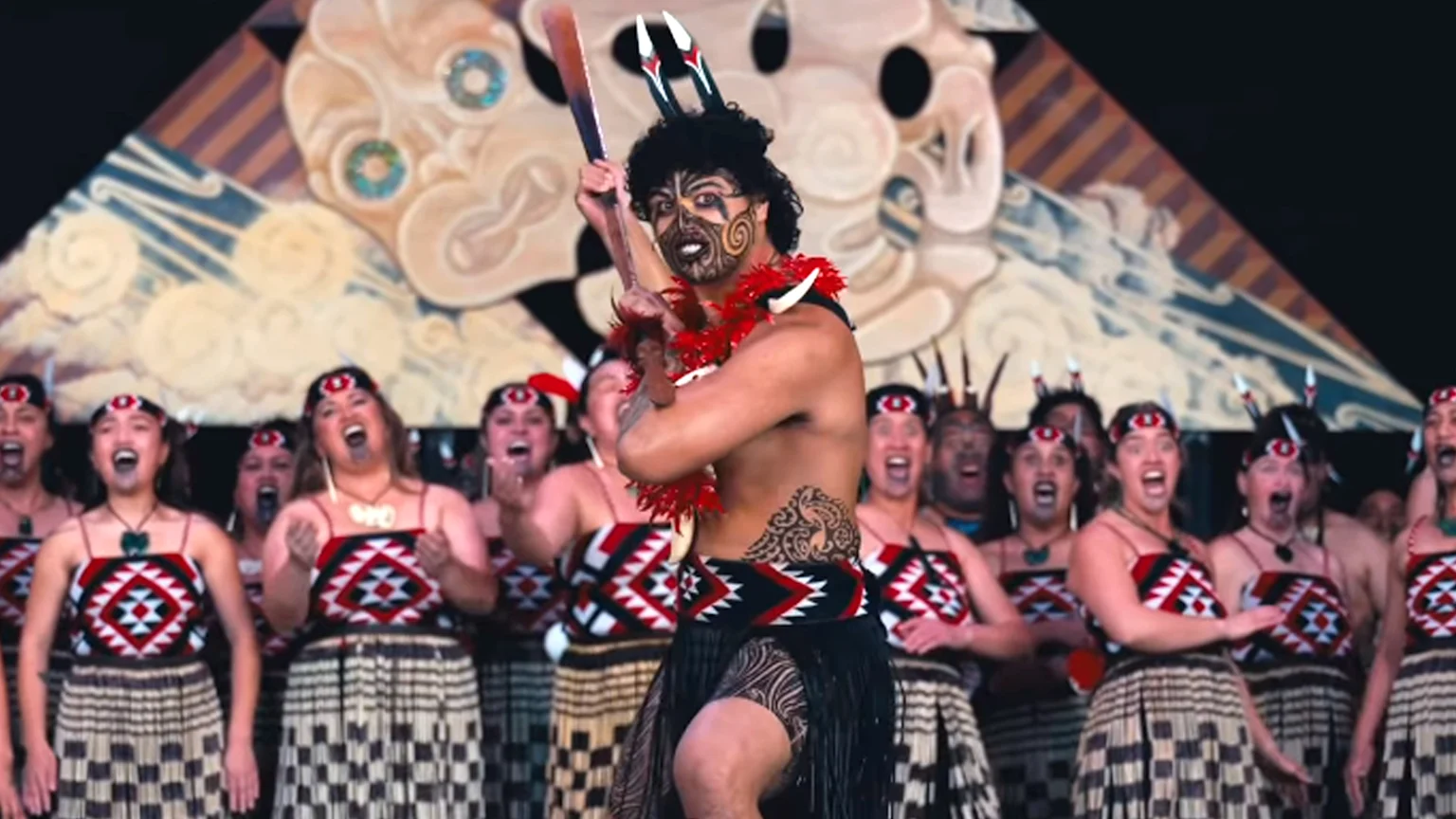A Maori man with face paint and a taiaha performs a haka in front of a group of women in traditional dress and a carved backdrop.