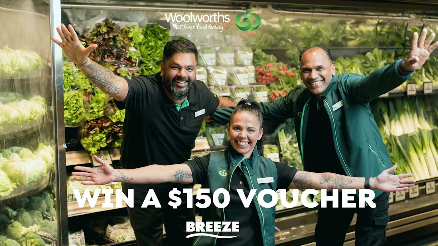 Three happy Woolworths employees in green uniforms, with arms outstretched, stand in a fresh produce aisle, surrounded by vegetables, with text "WIN A $150 VOUCHER" prominently displayed.