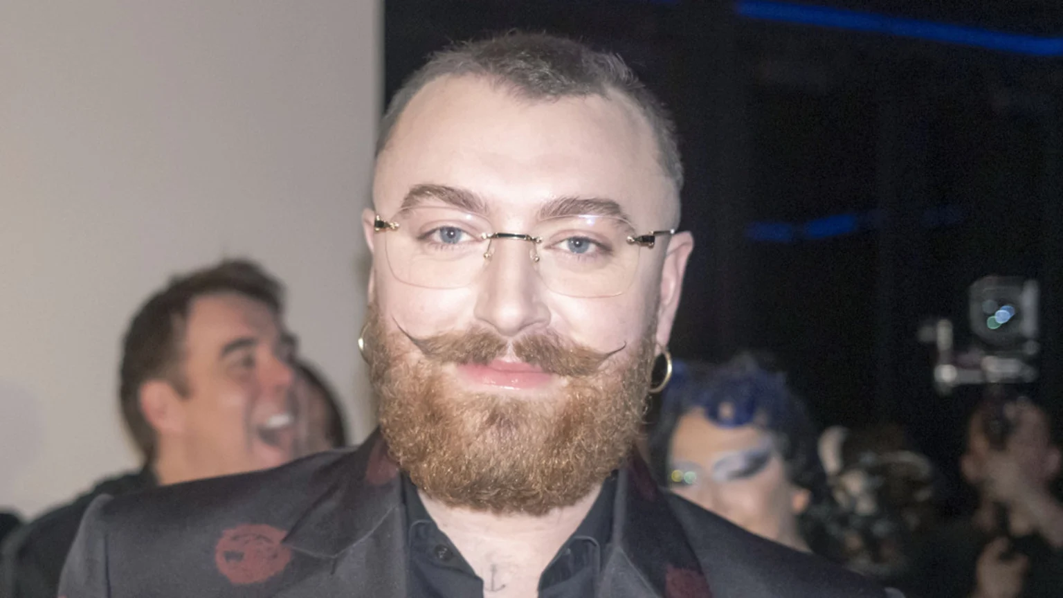 Sam Smith looks directly at the camera, sporting a ginger beard, stylized mustache, rimless glasses, and gold hoop earrings, wearing a dark patterned jacket.