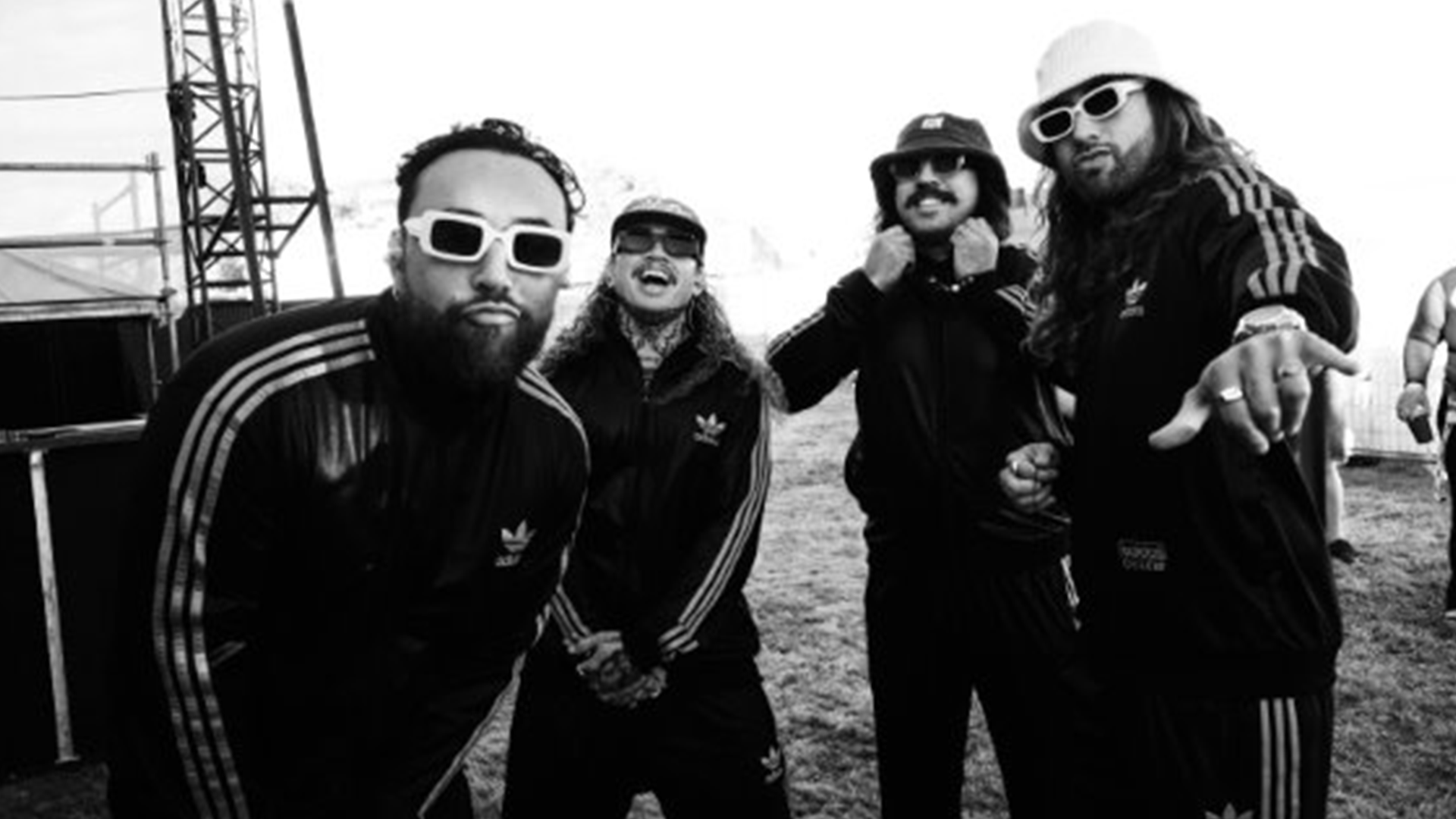 Four men in black Adidas tracksuits, sunglasses, and hats pose for the camera in a black and white outdoor shot.