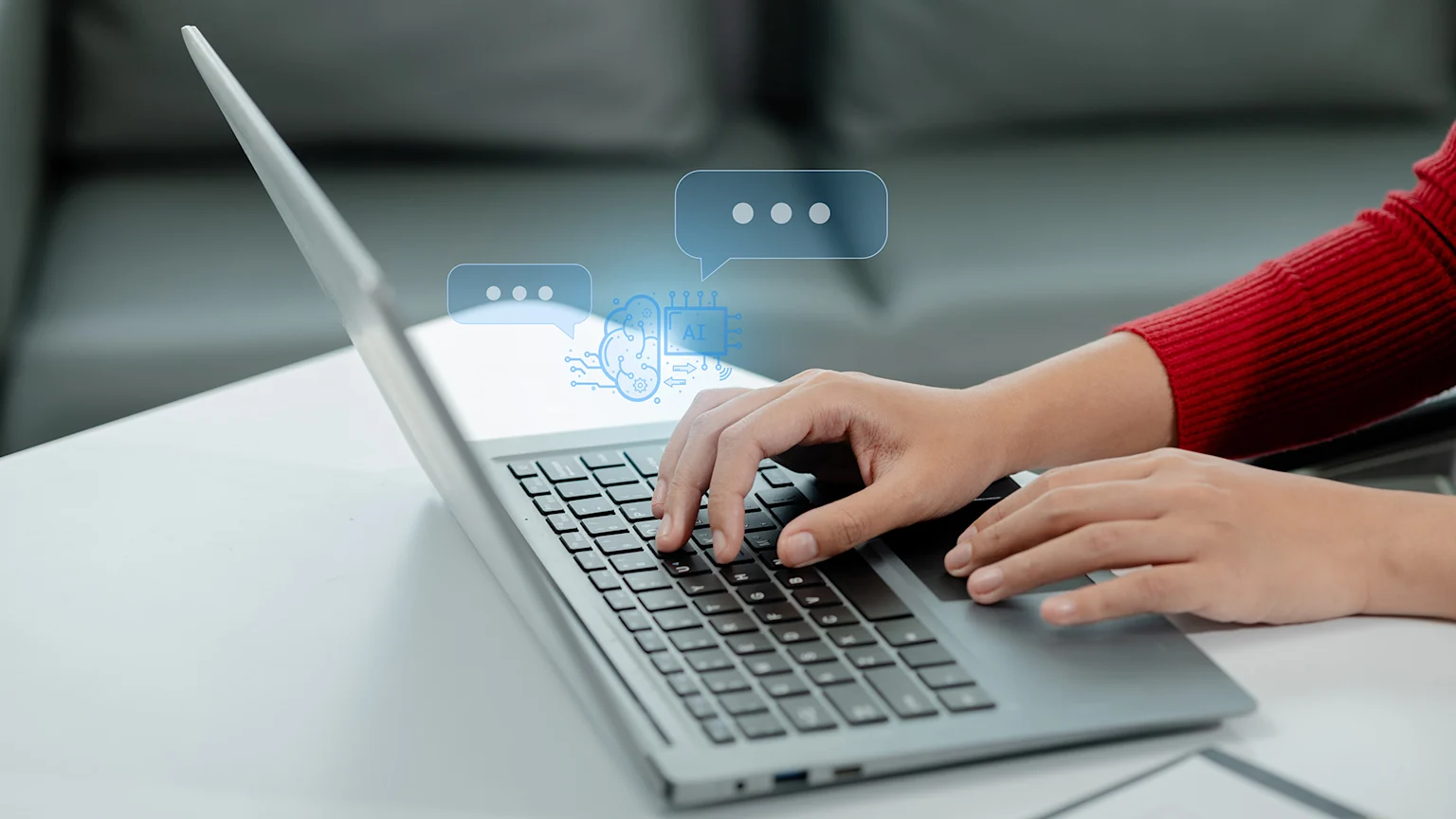 Close-up of a person's hands typing on a laptop keyboard, with blue AI-related icons and chat bubbles overlaid above the keyboard, on a white desk.