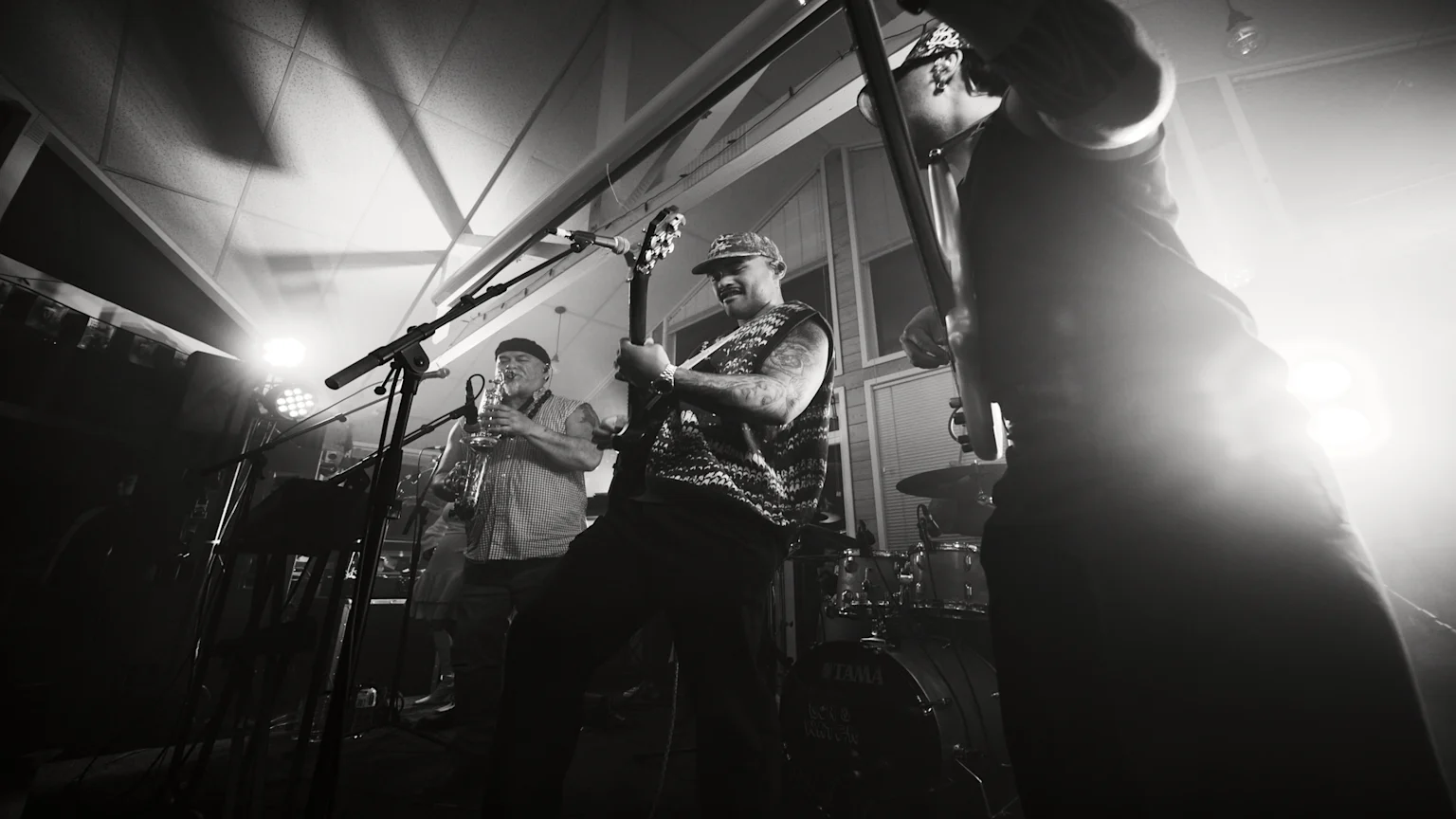 Black and white photo of the band Son & Water performing live on stage, with a saxophonist, a guitarist, and a bassist, under bright spotlights causing lens flare.