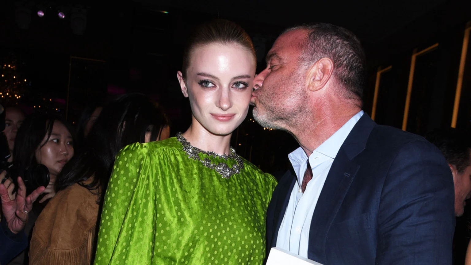 A smiling woman in a green polka-dot dress and a sparkly necklace receives a kiss on the cheek from a man with a beard.