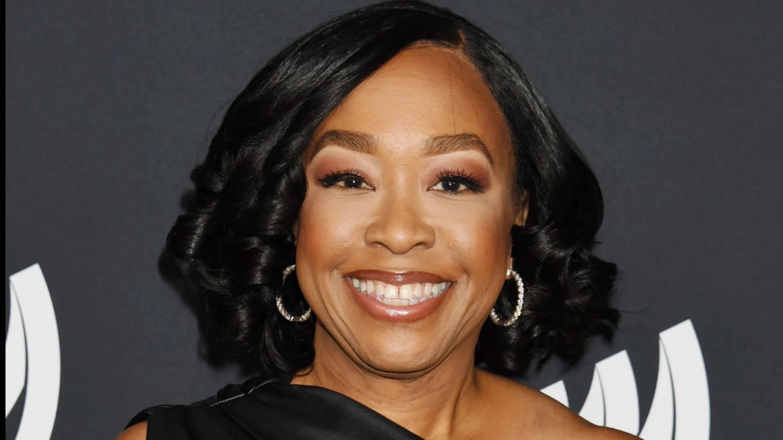 A woman with dark, styled hair and diamond hoop earrings smiles broadly, looking directly at the camera, against a dark background with a partial white logo.