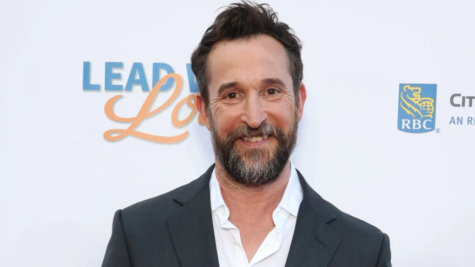 A smiling man with dark hair and a beard wears a dark suit jacket over an unbuttoned white shirt, against a white background with partial "LEAD," "Lo," and "RBC" logos.