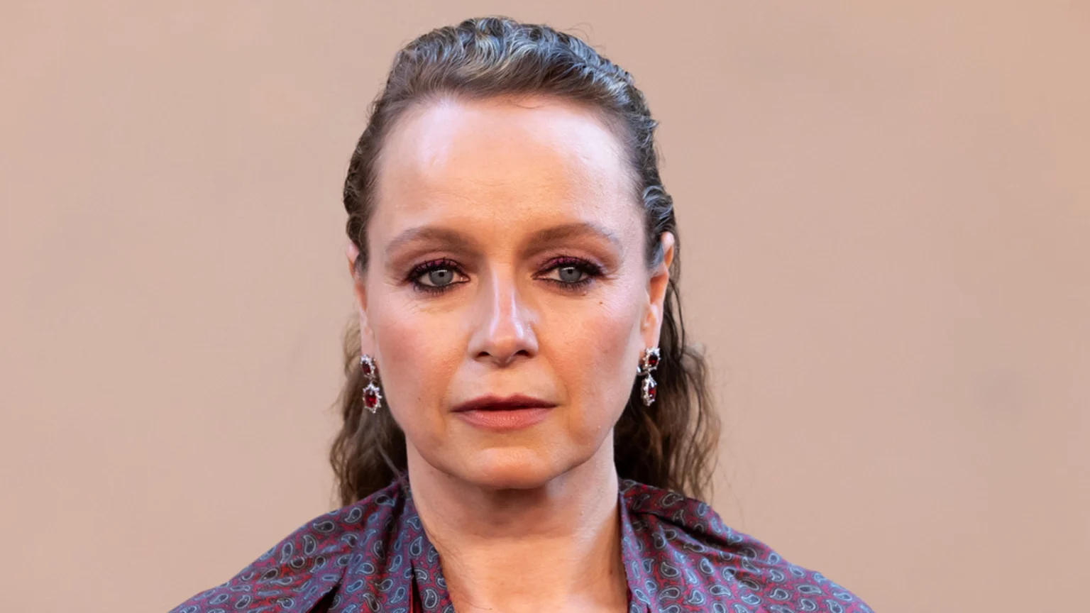 A woman with wet-look hair, pink eyeshadow, and red drop earrings looks directly at the camera. She wears a paisley patterned top against a plain beige background.