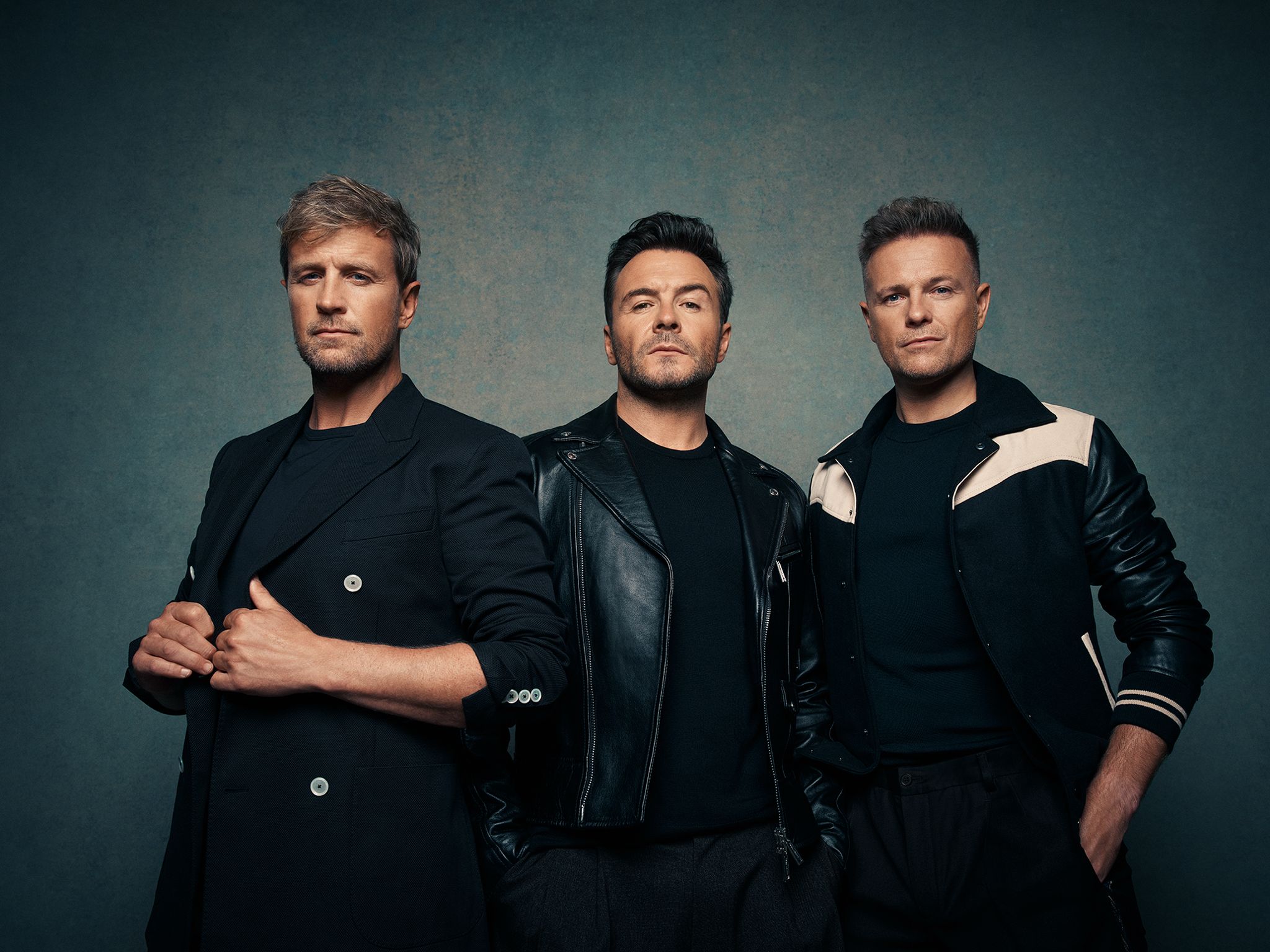 Three men, one in a suit jacket, one in a leather jacket, and one in a bomber jacket, stand side-by-side against a dark, textured background, looking directly at the camera.