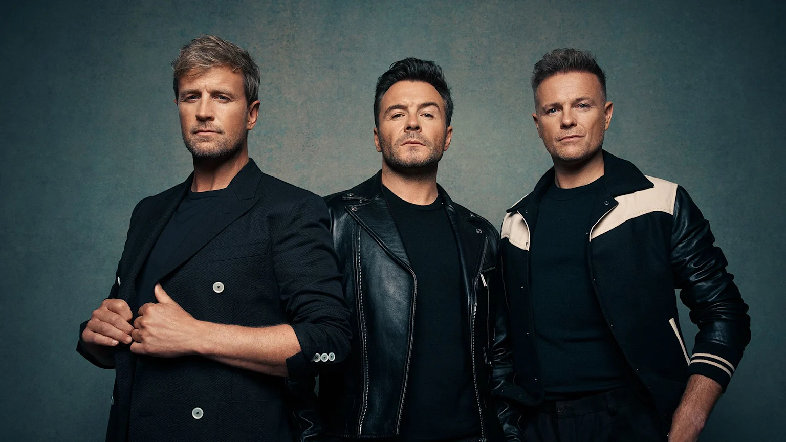 Three men, one in a suit jacket, one in a leather jacket, and one in a bomber jacket, stand side-by-side against a dark, textured background, looking directly at the camera.