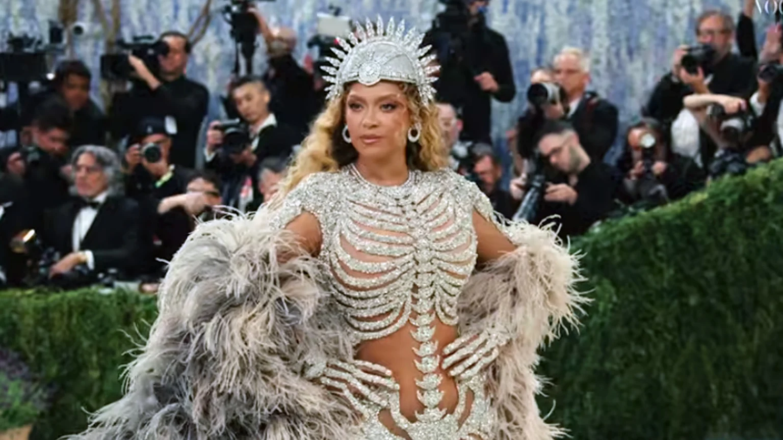Beyoncé wears a gem-encrusted gown designed like a skeleton, with feather accents and a crown, while posing at a formal event, surrounded by photographers.