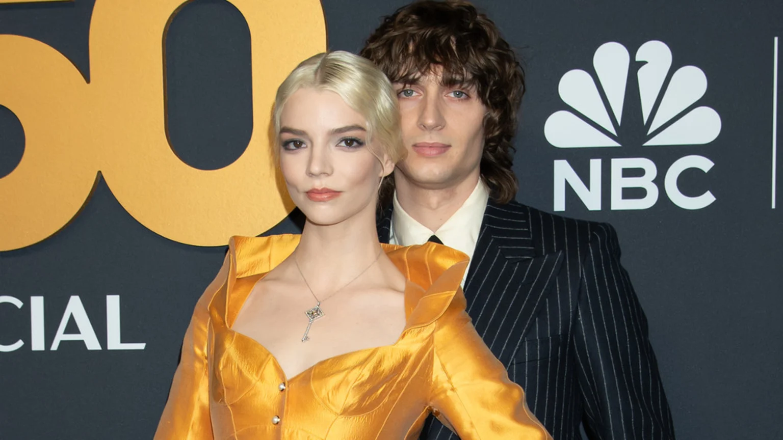 Anya Taylor-Joy in a gold dress and Malcolm McRae in a pinstripe suit pose together, with a golden "50" logo and the NBC logo visible in the background.