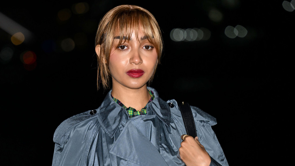 A young woman with light brown bangs and red lipstick wears a grey trench coat over a plaid shirt, looking directly at the camera with a dark, blurred background.