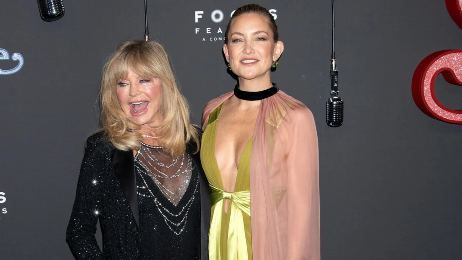 Goldie Hawn, in a black sequined top, smiles widely next to Kate Hudson, in a chartreuse and peach deep V-neck gown, against a dark background with microphones.