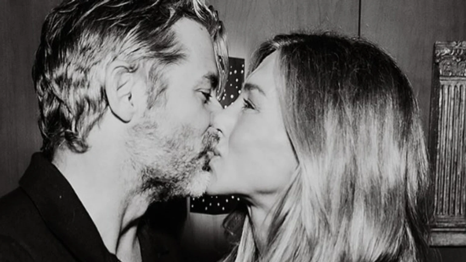 A black and white close-up photograph captures a man with short wavy hair and a beard and a woman with long hair in a tender kiss. Both are shown in profile with eyes closed, set against a dark indoor background featuring a fluted column and a spotted pattern.