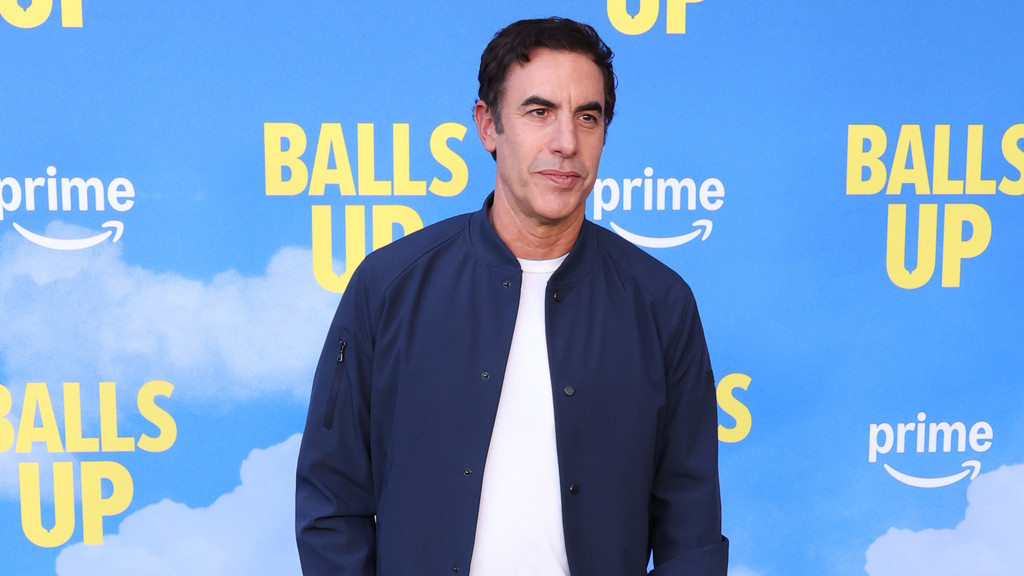 A man in a navy bomber jacket and white t-shirt stands in front of a blue backdrop with yellow "BALLS UP" text and white Amazon Prime logos.