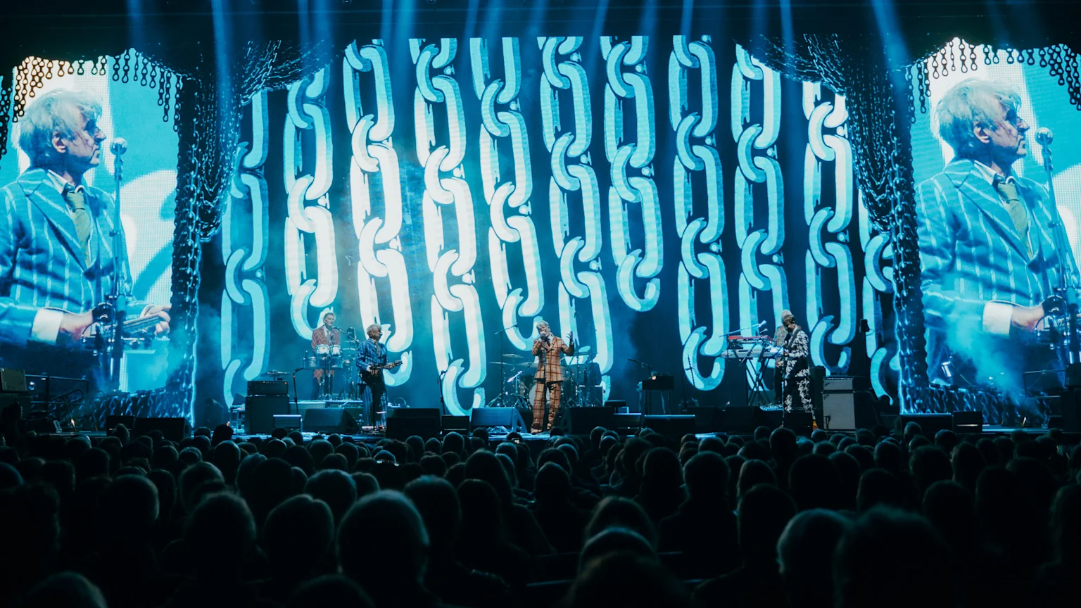 A band performs on a brightly lit concert stage with a large LED screen behind them showing a man in a striped suit and chain patterns, while an audience watches from below.