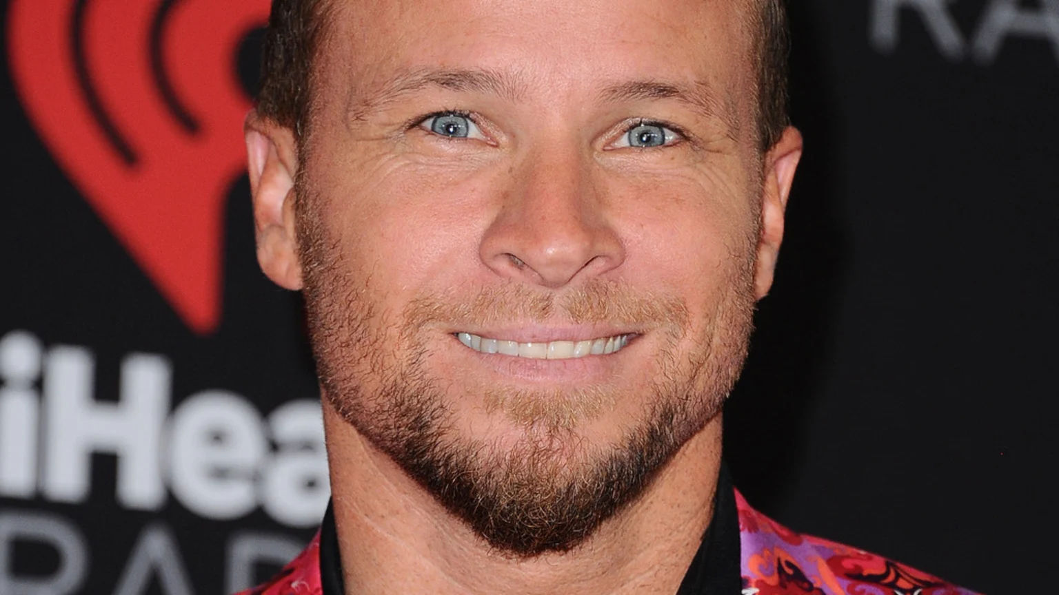 Close-up of a smiling man with blue eyes and a light brown beard, wearing a patterned shirt, with a red iHeartRadio logo in the background.