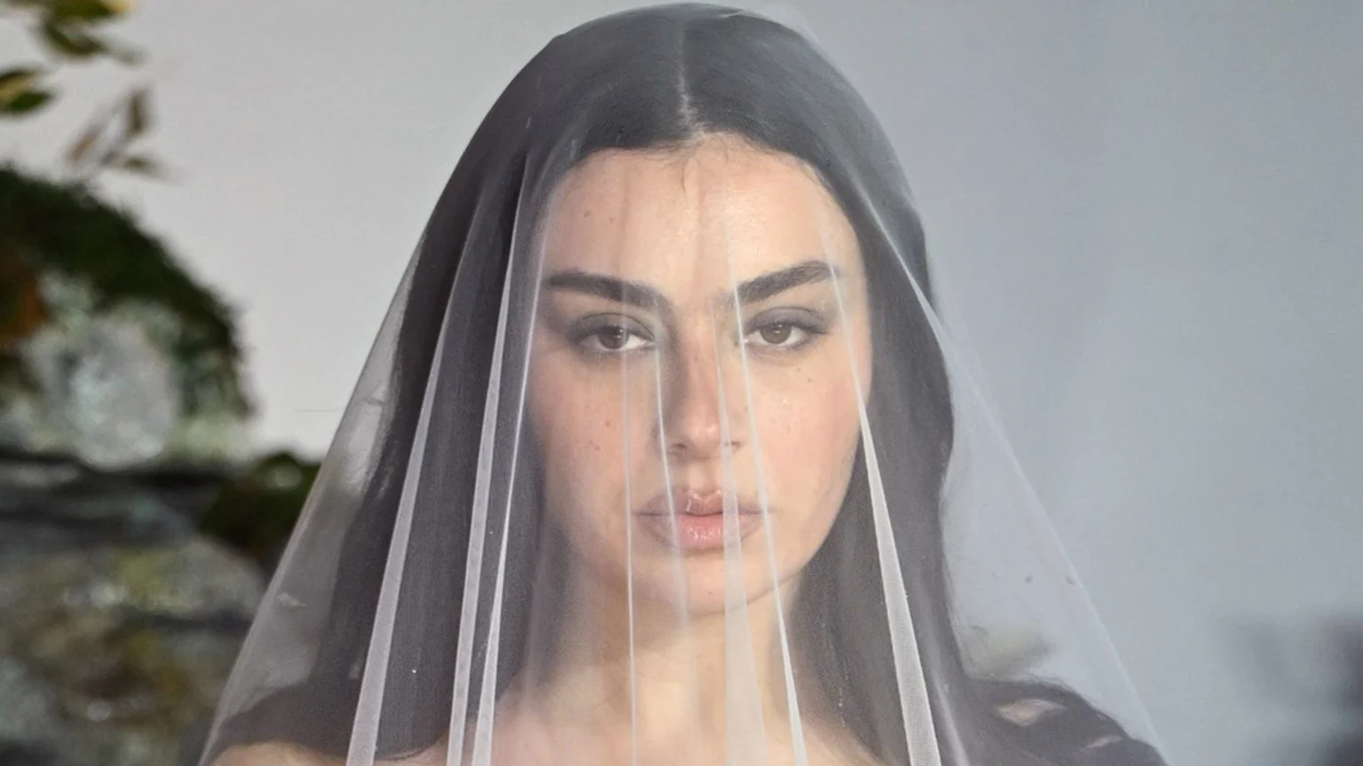 A woman with dark hair, prominent eye makeup, and glossy lips looks directly forward through a sheer white veil.
