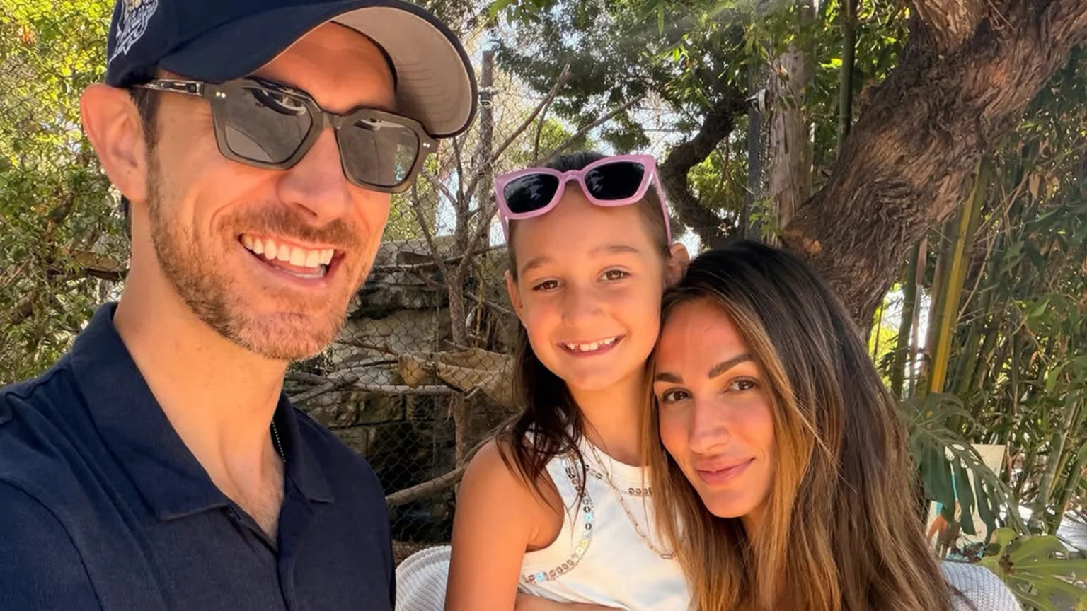 A smiling man in a cap and sunglasses takes a selfie with a girl wearing pink sunglasses on her head and a woman with long brown hair, all posing outdoors.
