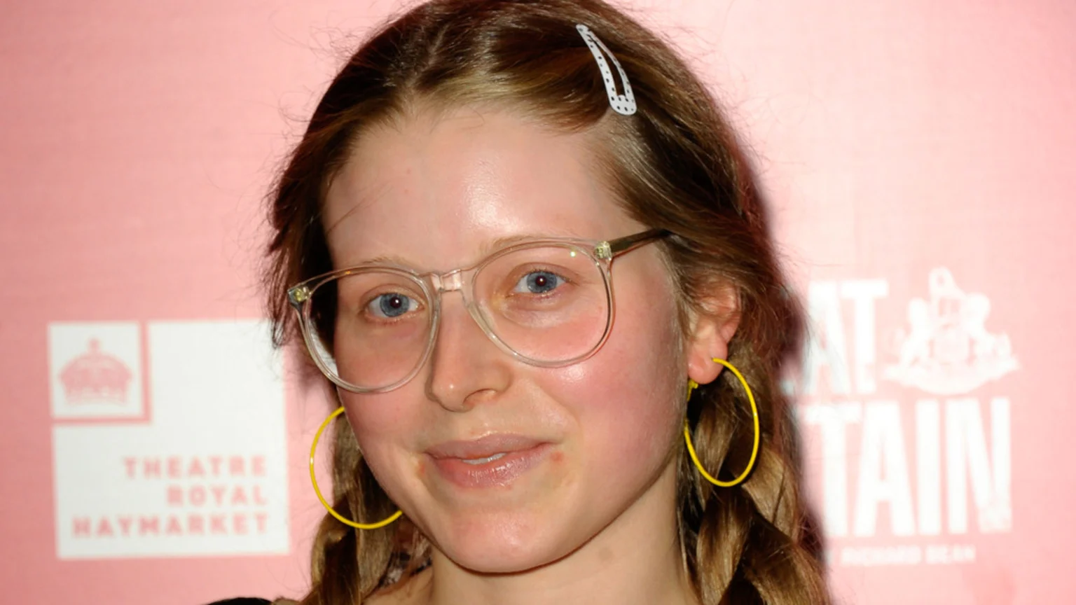 A smiling young woman with light brown braided hair, blue eyes, clear glasses, yellow hoop earrings, and a white polka-dot hair clip, against a pink background with blurred text.