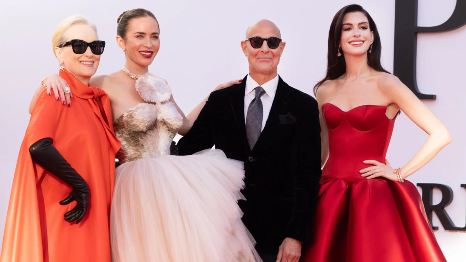 Meryl Streep in an orange cape, Emily Blunt in a light gown, Stanley Tucci in a dark suit, and Anne Hathaway in a red gown smile side-by-side at an event.
