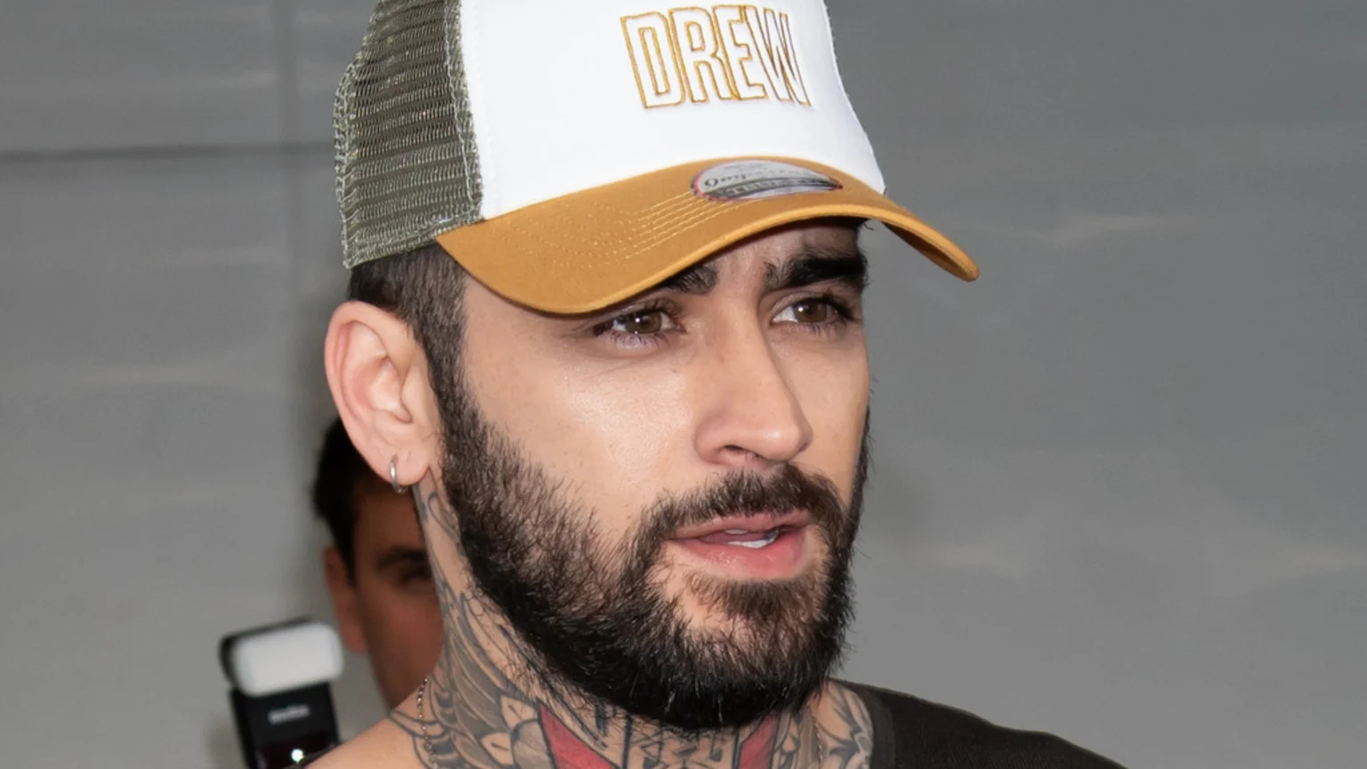 Close-up of Zayn Malik wearing a white and gold "DREW" trucker hat, with a beard, neck tattoos, and a silver hoop earring, looking towards the right.