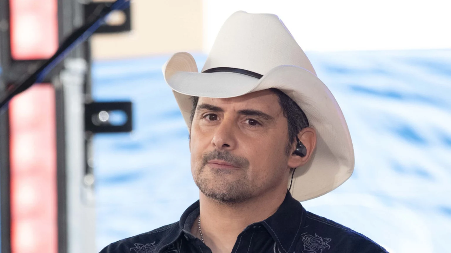 Brad Paisley, wearing a white cowboy hat, a dark patterned shirt, and an in-ear monitor, looks slightly left with a neutral expression.