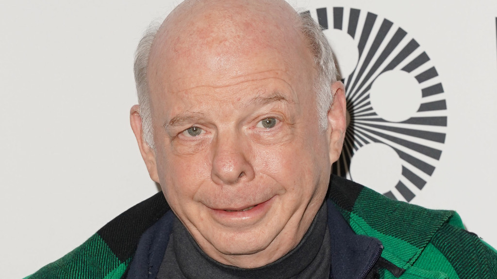 Close-up of actor Wallace Shawn smiling slightly, wearing a dark turtleneck and a green and black plaid jacket, with a black and white circular logo in the background.