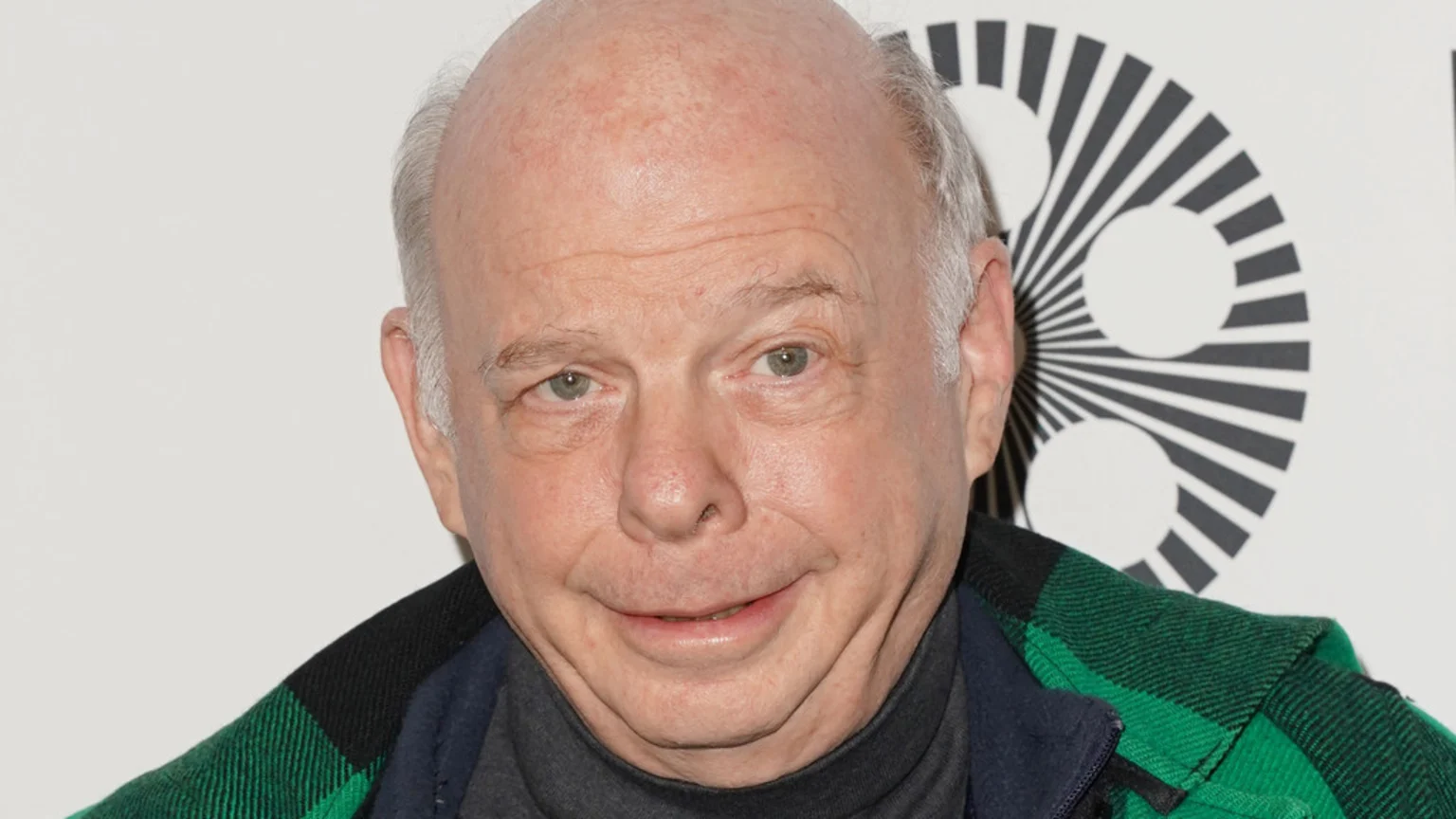 Close-up of actor Wallace Shawn smiling slightly, wearing a dark turtleneck and a green and black plaid jacket, with a black and white circular logo in the background.