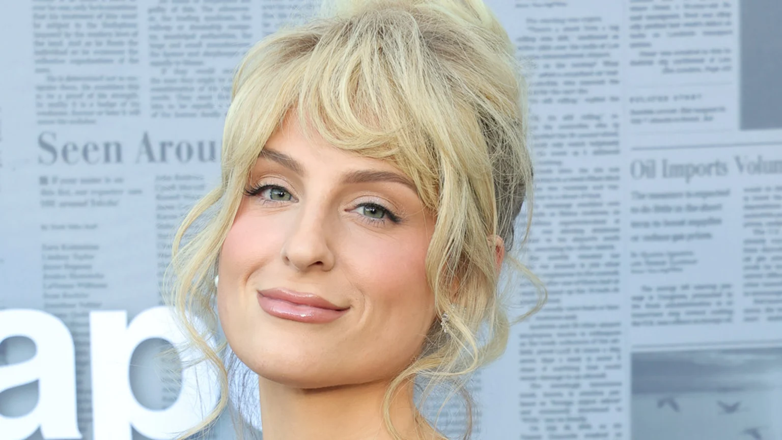 A blonde woman with bangs, light eye makeup, and glossy lips smiles slightly in a close-up portrait against a blurred background of newspaper text.