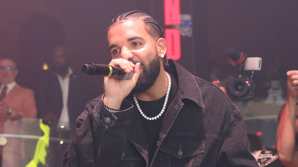 Drake, with braids and a beard, performs on stage holding a microphone, wearing a black denim jacket and a diamond chain.