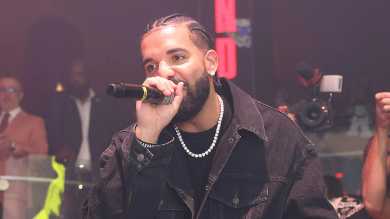 Drake, with braids and a beard, performs on stage holding a microphone, wearing a black denim jacket and a diamond chain.