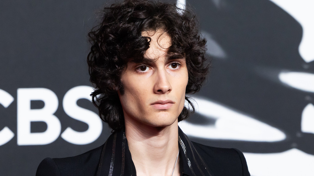 A person with dark curly hair and a serious expression, wearing a dark jacket and a silver necklace, is seen in a medium close-up against a dark background with a partially visible white 'CBS' logo.
