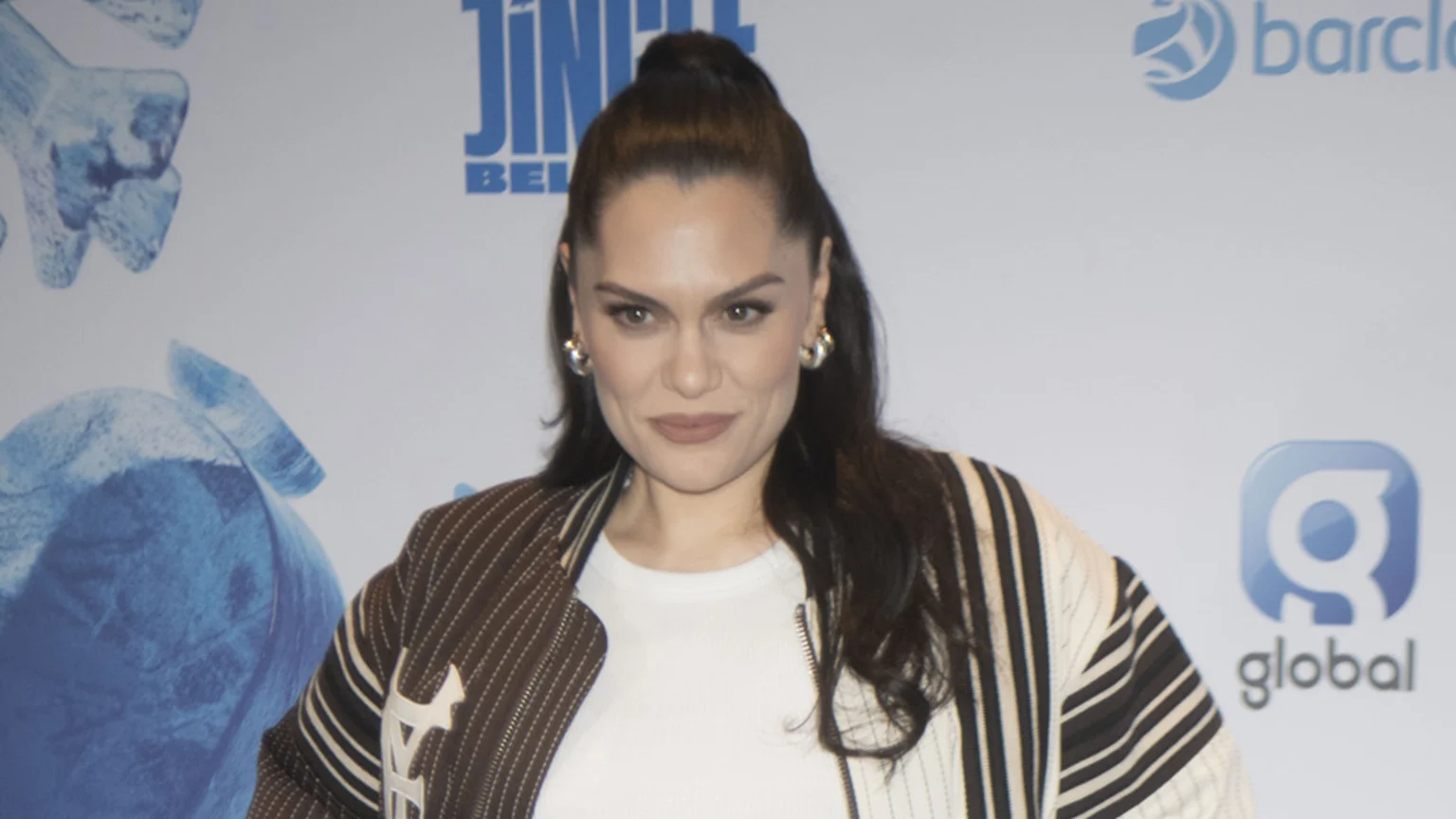Jessie J, with dark hair in a high ponytail and hoop earrings, looks towards the viewer, wearing a brown and white striped jacket over a white top, against a light background with sponsor logos.