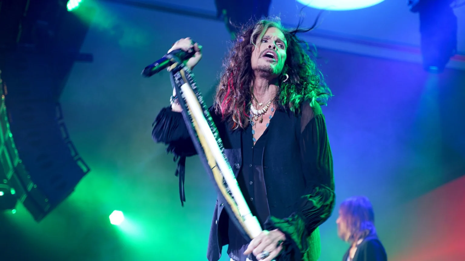 Steven Tyler, a male rock star with long curly hair and necklaces, sings into a microphone on stage, illuminated by green and blue lights, with another person visible in the background.