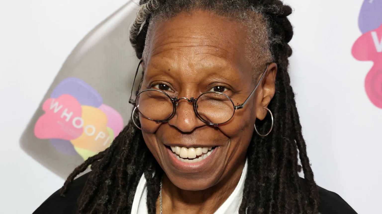 A close-up of Whoopi Goldberg smiling widely, wearing round glasses and hoop earrings, with her dreadlocked hair, against a blurry background with the word "WHOOPI".