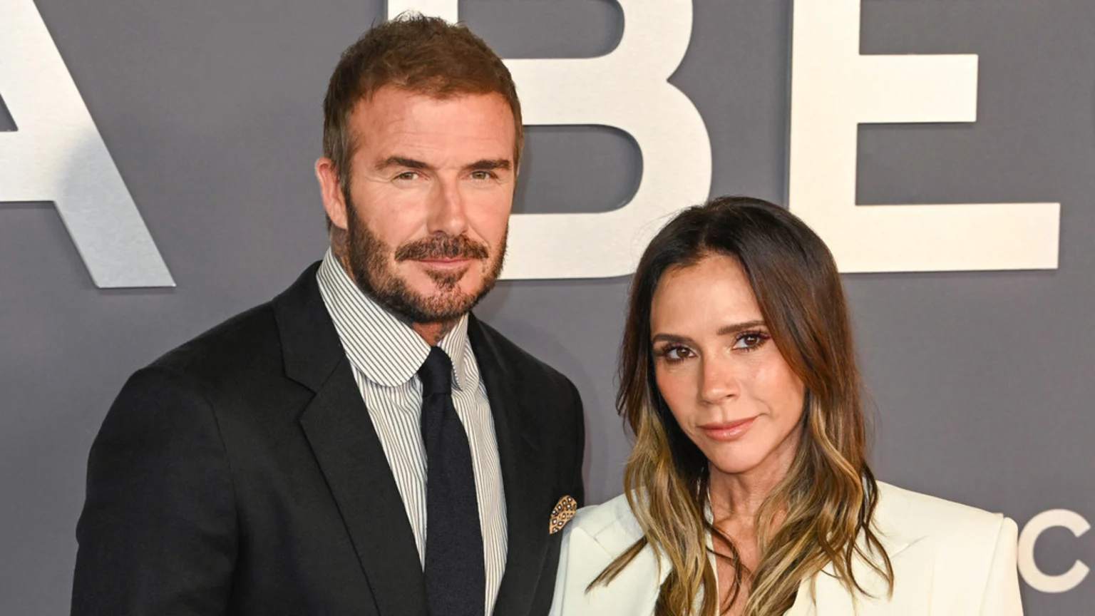 David and Victoria Beckham pose side-by-side, with David in a black suit and Victoria in a white blazer, against a grey background with large white letters.