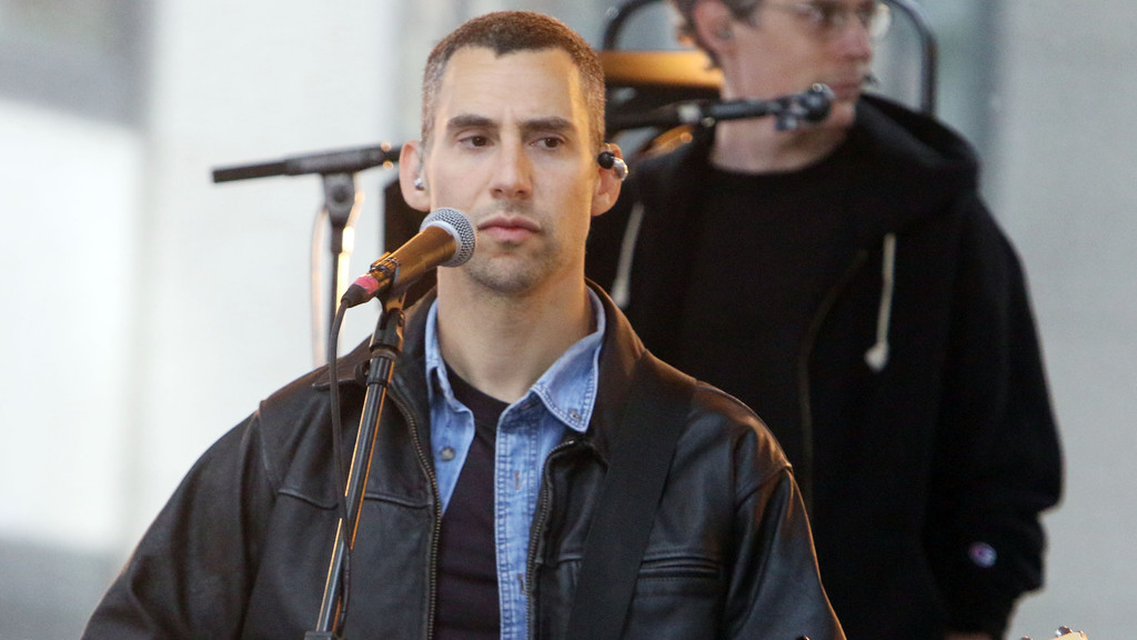 A man with short dark hair, wearing a black leather jacket over a denim shirt, stands at a microphone with in-ear monitors, looking forward with a blurry figure behind him.
