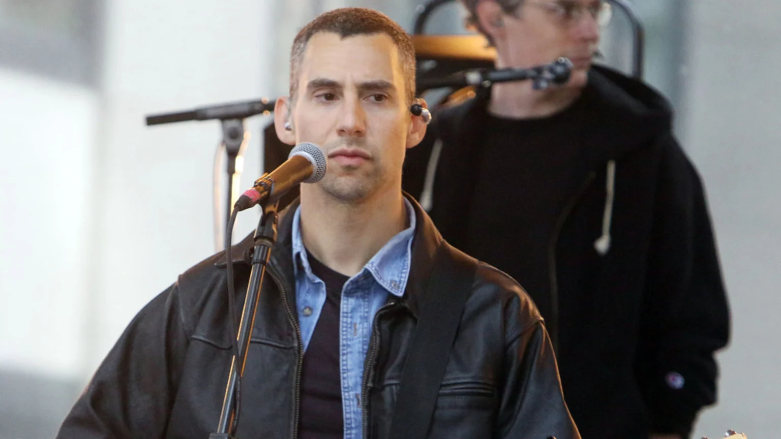 A man with short dark hair, wearing a black leather jacket over a denim shirt, stands at a microphone with in-ear monitors, looking forward with a blurry figure behind him.