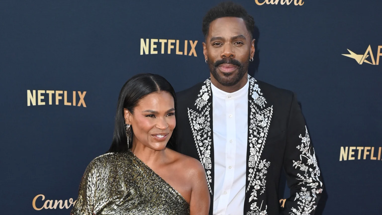 Colman Domingo, in an embroidered black blazer, and Nia Long, in a gold one-shoulder top, pose for a photo on a blue backdrop with Netflix logos.